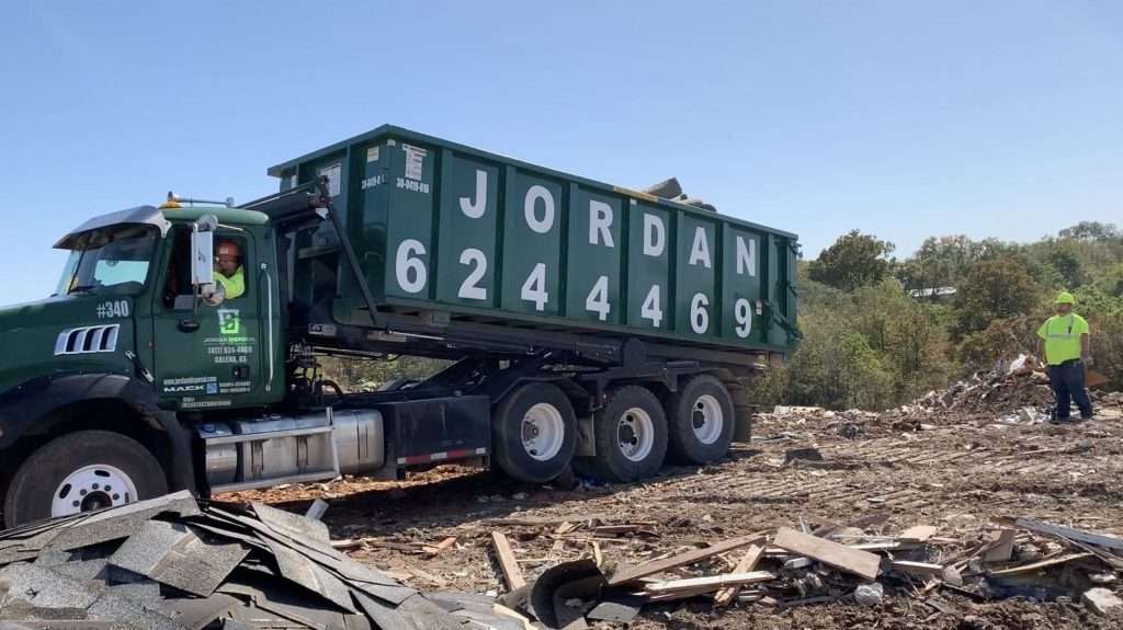 A large green dumpster truck labeled JORDAN and a phone number unloads debris at a construction site. Two workers in safety vests and helmets stand nearby on a sunny day.