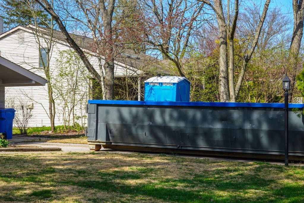 Empty green dumpster with a blue portable potty are seen in a driveway in front of a small house in a residential neighborhood.
